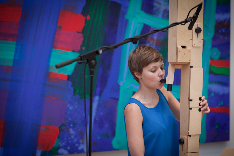 A female student performing a wooden instrument in front of an abstract painting.